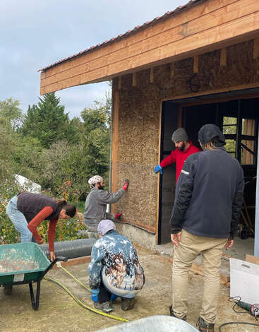 Five people are working together on the exterior wall of a wooden building, measuring, installing straw, and using tools, showing teamwork and construction skills outdoors.