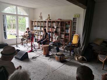 Three musicians perform in a cozy living room, surrounded by bookshelves and natural light. An audience listens attentively, seated comfortably in armchairs and on the floor.