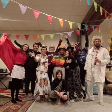A joyful group of people in creative costumes pose indoors beneath colorful bunting, celebrating together with bright smiles, playful gestures, and festive decorations in the background.