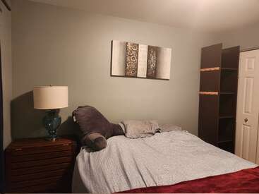 Une chambre douillette avec un grand lit, des murs gris, une lampe en verre bleu posée sur une table de chevet en bois, une décoration murale abstraite et des étagères marron vides à côté d'une porte blanche.