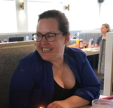 Une femme portant des lunettes et une chemise bleue sourit joyeusement, assise à une table de restaurant. À l'arrière-plan, on aperçoit d'autres clients et des condiments sur les tables.