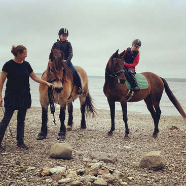 Das Bild zeigt zwei Kinder, die auf Pferden an einem felsigen Strand reiten, während ein Erwachsener in der Nähe steht, vor einem ruhigen Hintergrund aus Wasser und bedecktem Himmel.