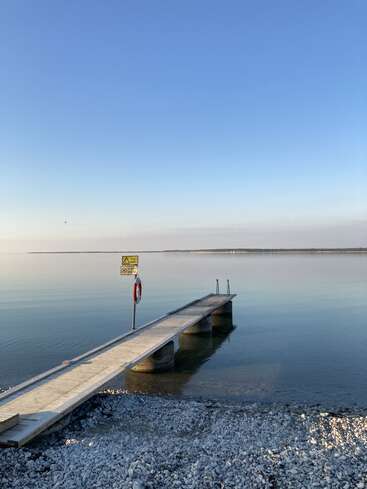 Das Bild zeigt eine ruhige Seeszene mit einem langen hölzernen Steg, der ins Wasser ragt, mit einem gelben Schild und einer Rettungsweste vor einem klaren blauen Himmel.