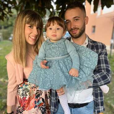 The image depicts a family of three posing for a photo, with the parents holding their young child in a blue dress, standing outdoors in front of a building.