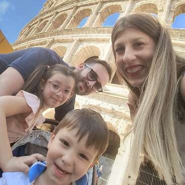 The image shows a family of four posing for a selfie in front of the Colosseum in Rome, Italy, with a bright blue sky in the background.
