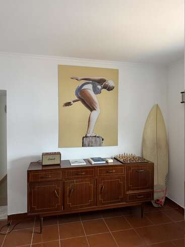 Essa imagem mostra um aparador de madeira do meio do século com livros, um jogo de xadrez e um alto-falante. Acima dele, há um quadro grande de um mergulhador. Uma prancha de surfe está ali perto.
