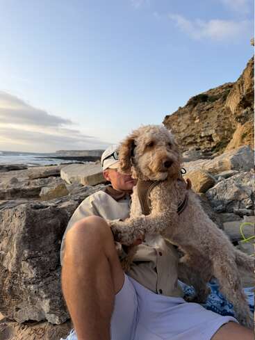 Uma pessoa está sentada numa praia rochosa, usando óculos escuros e um boné, abraçando carinhosamente um cachorro fofinho. O mar e as falésias criam um cenário sereno e tranquilo ao pôr do sol.