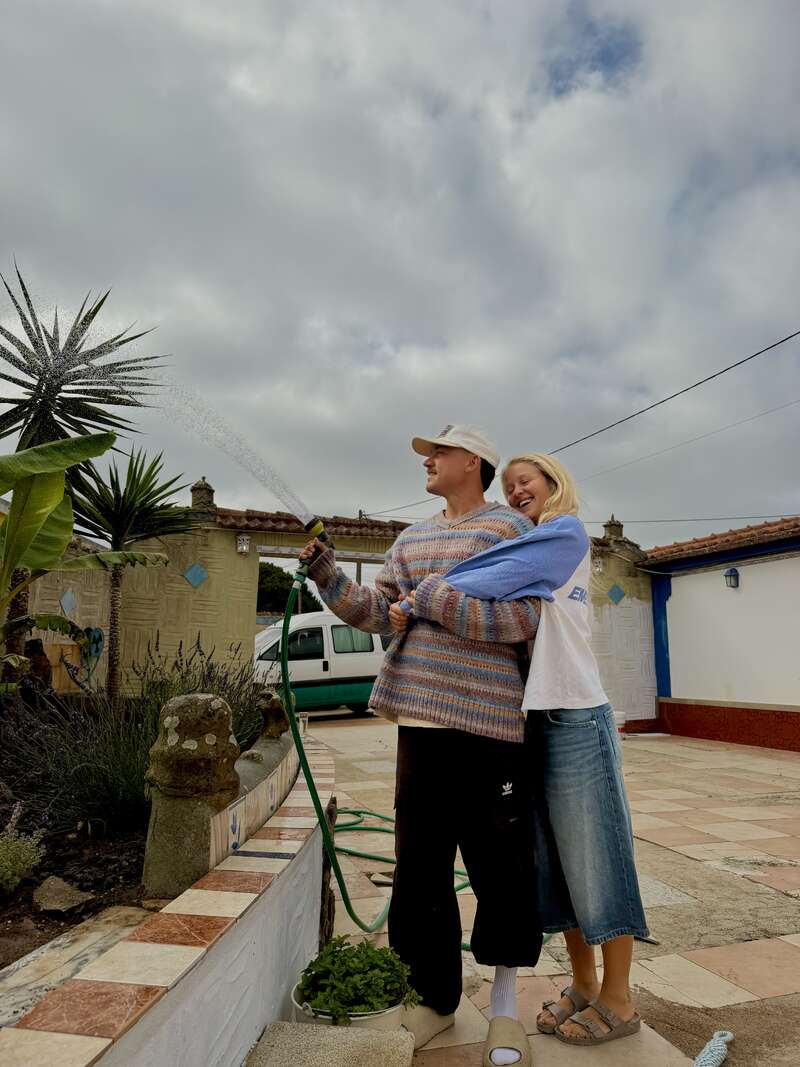 Um casal alegre rega as plantas num quintal ensolarado. O homem segura a mangueira enquanto a mulher o abraça por trás; os dois sorriem e aproveitam o momento juntos.
