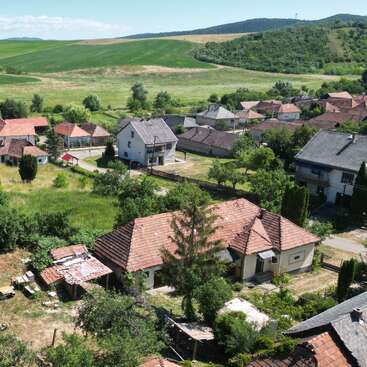 Cette image montre un petit village rural avec des maisons aux toits rouges, des jardins verts, des arbres et des collines ondulantes en arrière-plan sous un ciel bleu clair.