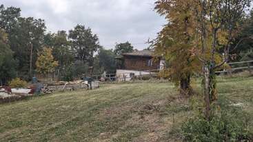 A rustic wooden house sits on a grassy hill, surrounded by autumn trees, scattered construction materials, and a wooden fence. The sky is cloudy and gray.