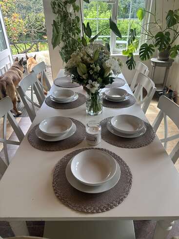 Une table à manger joliment dressée présente de la vaisselle blanche, des sets de table tissés, une composition florale et une bougie. Deux chiens se tiennent près d'une porte ouverte, avec de la verdure à l'extérieur.