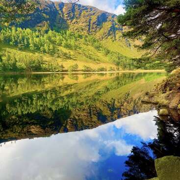 Un paisaje sereno con un lago cristalino en el que se reflejan laderas de un verde vibrante, árboles imponentes, orillas rocosas y un cielo azul brillante adornado con esponjosas nubes blancas.