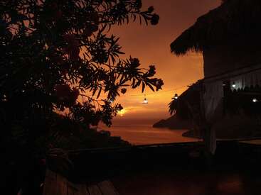 The image depicts a serene sunset scene, featuring a house with a thatched roof, a tree, and a body of water, all bathed in the warm glow of the setting sun.