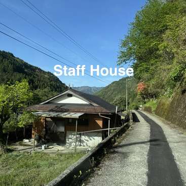 Ein kleines Haus mit der Aufschrift „Staff House“ steht an einer schmalen asphaltierten Straße, umgeben von üppig grünen Bäumen und Bergen unter einem strahlend blauen Himmel. Eine friedliche Atmosphäre.