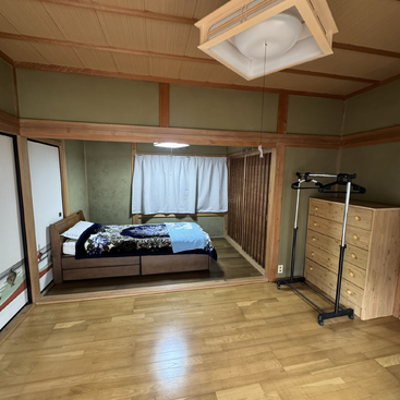 This is a spacious, traditional Japanese-style bedroom with tatami mats, wooden flooring, a single bed, dresser, clothes rack, and large window with white curtains.