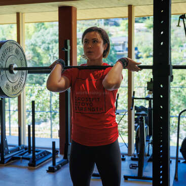 A woman in a red "CrossFit Otoyo Strength" shirt is lifting a barbell indoors at a gym, focused and determined, surrounded by gym equipment.