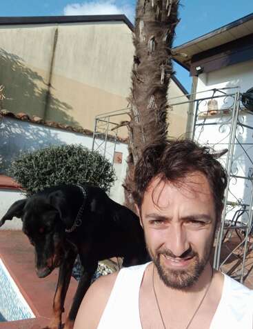 La imagen muestra a un hombre de pelo corto y camisa blanca, de pie frente a un fondo gris, con una expresión neutra en el rostro.