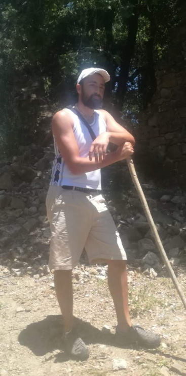 La imagen muestra a un hombre de pie frente a un árbol, vestido con camisa blanca y pantalones oscuros, con expresión seria en el rostro. Lleva un palo en la mano derecha.
