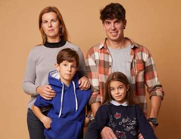 Una familia de cuatro miembros posa ante un fondo beige: la madre y el padre están de pie detrás de su hijo y su hija, y todos lucen expresiones tranquilas, relajadas y amables.