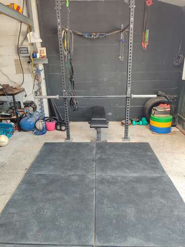En esta imagen se ve un gimnasio doméstico instalado en un garaje, con un soporte para press de banca, discos de pesas, bandas elásticas, equipo de tiro con arco, suelo de goma y diversas herramientas.