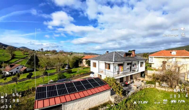 Una casa blanca de dos plantas se alza en un pintoresco paisaje rural, rodeada de vegetación. Los paneles solares se alzan sobre un edificio cercano de tejado rojo bajo un cielo de un azul intenso.
