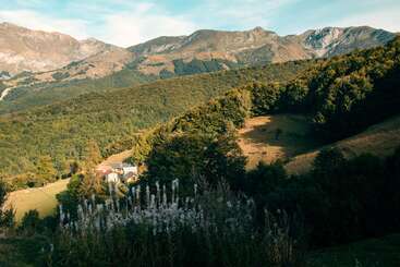 A picturesque mountain landscape features lush green hills, dense forests, a peaceful valley, and a small village nestled among the trees, under a bright sky.