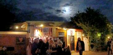 Un groupe animé se rassemble à l'extérieur d'un bar rustique chaleureusement éclairé la nuit. La lune se faufile à travers les nuages, jetant une lueur mystérieuse et atmosphérique sur la scène festive.