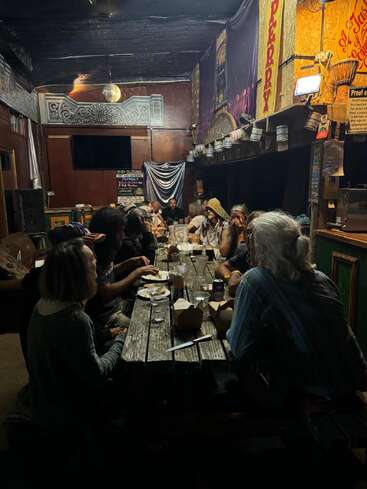 Un groupe de personnes se réunit autour d'une table en bois rustique dans une pièce éclectique faiblement éclairée, partageant nourriture et conversation, créant ainsi une atmosphère chaleureuse et communautaire.