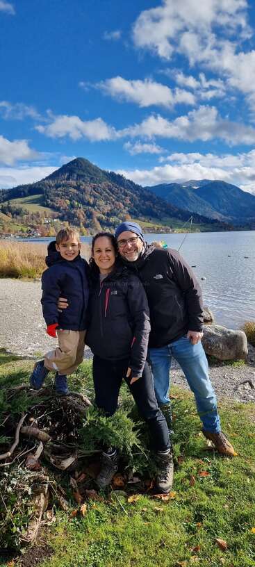 A smiling family of three poses outdoors by a scenic lake, surrounded by mountains, green grass, and a beautiful blue sky with fluffy clouds.