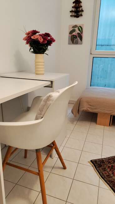 A cozy, minimalist room featuring a white desk with a vase of pink flowers, comfortable white chair, single bed, wall art, window, and patterned rug.