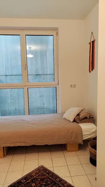 A minimal bedroom with a single bed, beige bedding, large window, wall hanging with colorful ribbons, a woven basket, and a patterned carpet on tiled floor.