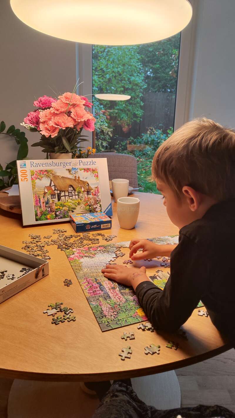 A child concentrates on completing a colorful jigsaw puzzle at a cozy table, surrounded by flowers, mugs, and natural light from a large window.