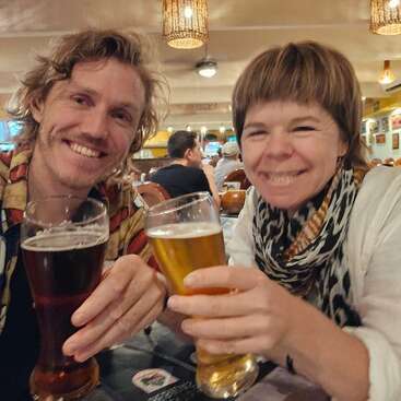 The image depicts a man and woman sitting at a table, each holding a glass of beer, smiling at the camera, in a restaurant or bar setting.