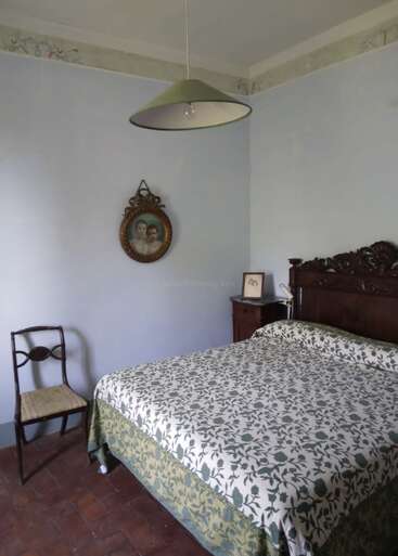 This image shows a simple, vintage bedroom with a patterned bedspread, wooden chair, ornate portrait, wooden headboard, nightstand, and a single green lampshade overhead.