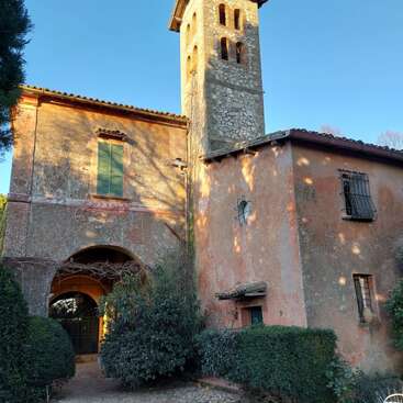 A rustic stone building with a tall bell tower, green shuttered windows, arched entrance, and climbing ivy, bathed in sunlight against a clear blue sky.