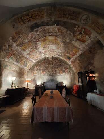This image shows a dimly lit, historic dining room with a long table, ornate chairs, antique furniture, and a stunning frescoed arched ceiling depicting religious scenes.