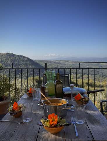 Une table rustique dressée pour un repas en plein air surplombe des collines ondulantes sous un ciel bleu clair. Des salades colorées, des soupes, des boissons et des fleurs éclatantes décorent la scène.