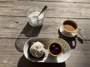 Une table rustique en bois présente du café, un bol de sucre en verre transparent, un brownie recouvert de crème et un petit plat de baies mélangées, le tout baigné par la lumière du soleil.