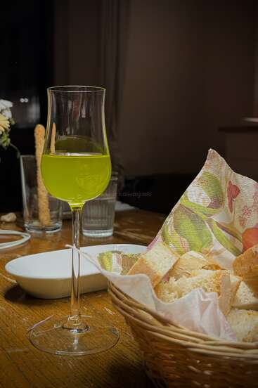 Un verre de liqueur vert-jaune est posé sur une table en bois à côté d'une corbeille de pain, d'une serviette, d'un bol blanc et de verres, créant ainsi une scène de repas confortable.
