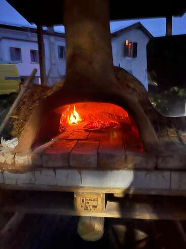 A rustic outdoor wood-fired oven glows warmly at dusk, with a pizza baking inside. The scene is cozy, inviting, and perfect for homemade meals.