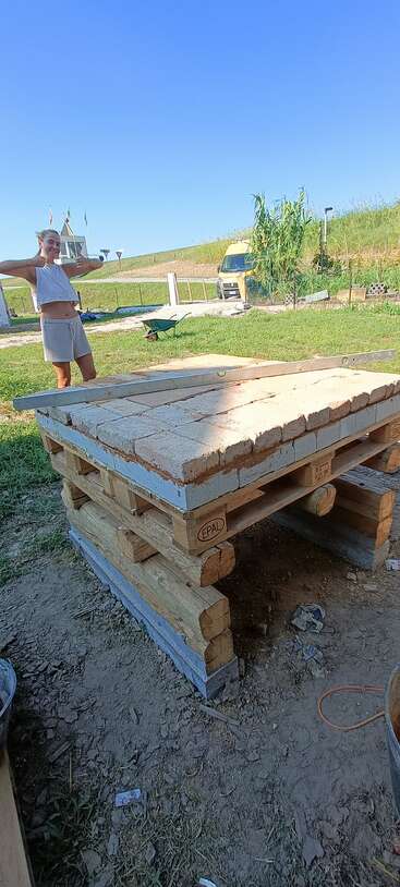 A person smiles and gives two thumbs up near a homemade structure built from bricks, pallets, and logs, outside on a sunny day with blue sky.