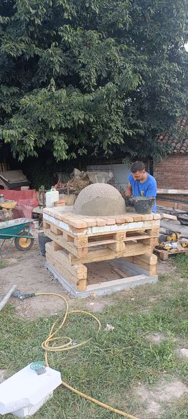 A man works outdoors on building a brick and cement dome structure, possibly an oven, surrounded by tools, wood pallets, and lush green trees.