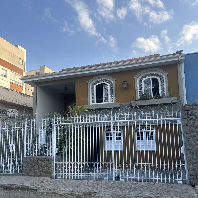 This image shows a two-story house with decorative windows, a brown facade, and a white metal fence. Barbed wire tops the fence, and plants are visible.