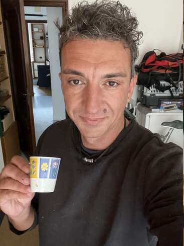 A person with short, curly, gray-streaked hair smiles, holding a small, colorful floral cup. They stand indoors, with shelves and bags visible in the background.