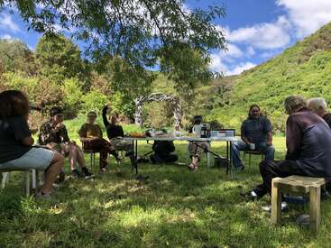 Un groupe de personnes est assis en plein air autour d'une table, dégustant des plats et discutant sous un arbre dans un paysage pittoresque, vert et vallonné, par une journée ensoleillée.