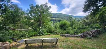 This peaceful outdoor scene features a hammock, a bench, and a chair amidst lush greenery, overlooking forested hills under a partly cloudy blue sky. Logs lie nearby.
