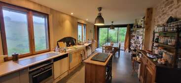 This image shows a cozy, rustic kitchen with wooden cabinets, large windows, a kitchen island, shelves of jars, and a dining area overlooking greenery.