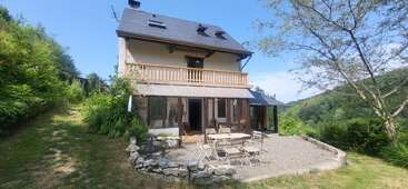 A charming three-story house with a balcony and glass doors, surrounded by lush greenery. Outdoor patio features a table and chairs, overlooking scenic, tree-covered hills.