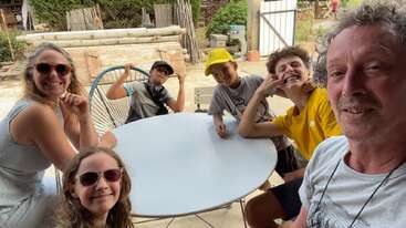 A group of six people, including adults and children, smile and pose around a white outdoor table. The background features yard equipment, wood, and greenery.