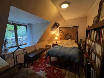 A cozy attic bedroom with slanted ceilings, single bed, bookshelf, tapestry wall art, red patterned rug, lamp, window seat, and warm, inviting lighting.